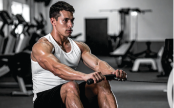 Boost It Naturally: Top 12 Natural Testosterone Boosters Muscular man in a white tank top rows a cable handle in a gym, focused on the exercise.
