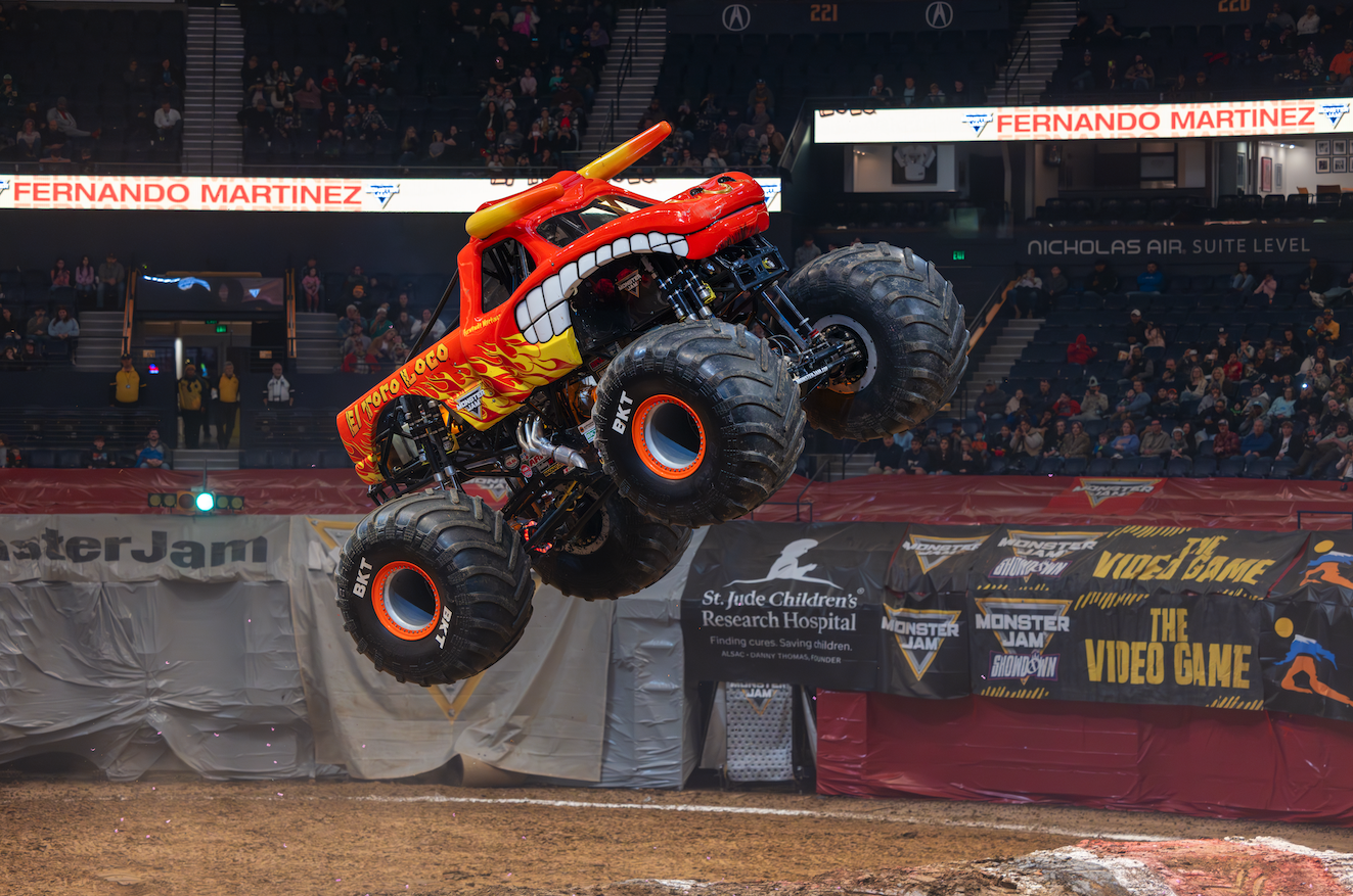 Orange monster truck with flaming design soaring mid-air over the dirt arena as spectators watch from stands.
