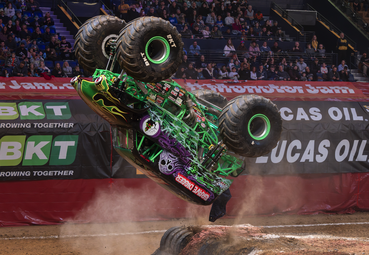 Monster truck in mid-air flip during a stadium arena show, green frame with purple skull artwork and large off-road tires; dusty dirt track and cheering crowd in background.