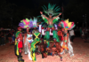 Out With Scott – By Scott Holland Carnival performers in colorful feathered costumes pose at night, central dancer on a pedestal with a large green headdress.
