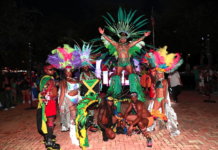 Carnival performers in colorful feathered costumes pose at night, central dancer on a pedestal with a large green headdress.