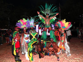 Out With Scott – By Scott Holland Carnival performers in colorful feathered costumes pose at night, central dancer on a pedestal with a large green headdress.
