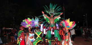 Carnival performers in colorful feathered costumes pose at night, central dancer on a pedestal with a large green headdress.