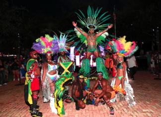 Carnival performers in colorful feathered costumes pose at night, central dancer on a pedestal with a large green headdress.