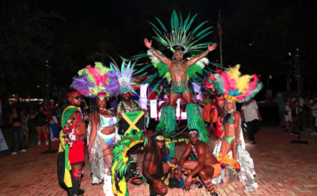 Out With Scott – By Scott Holland Carnival performers in colorful feathered costumes pose at night, central dancer on a pedestal with a large green headdress.