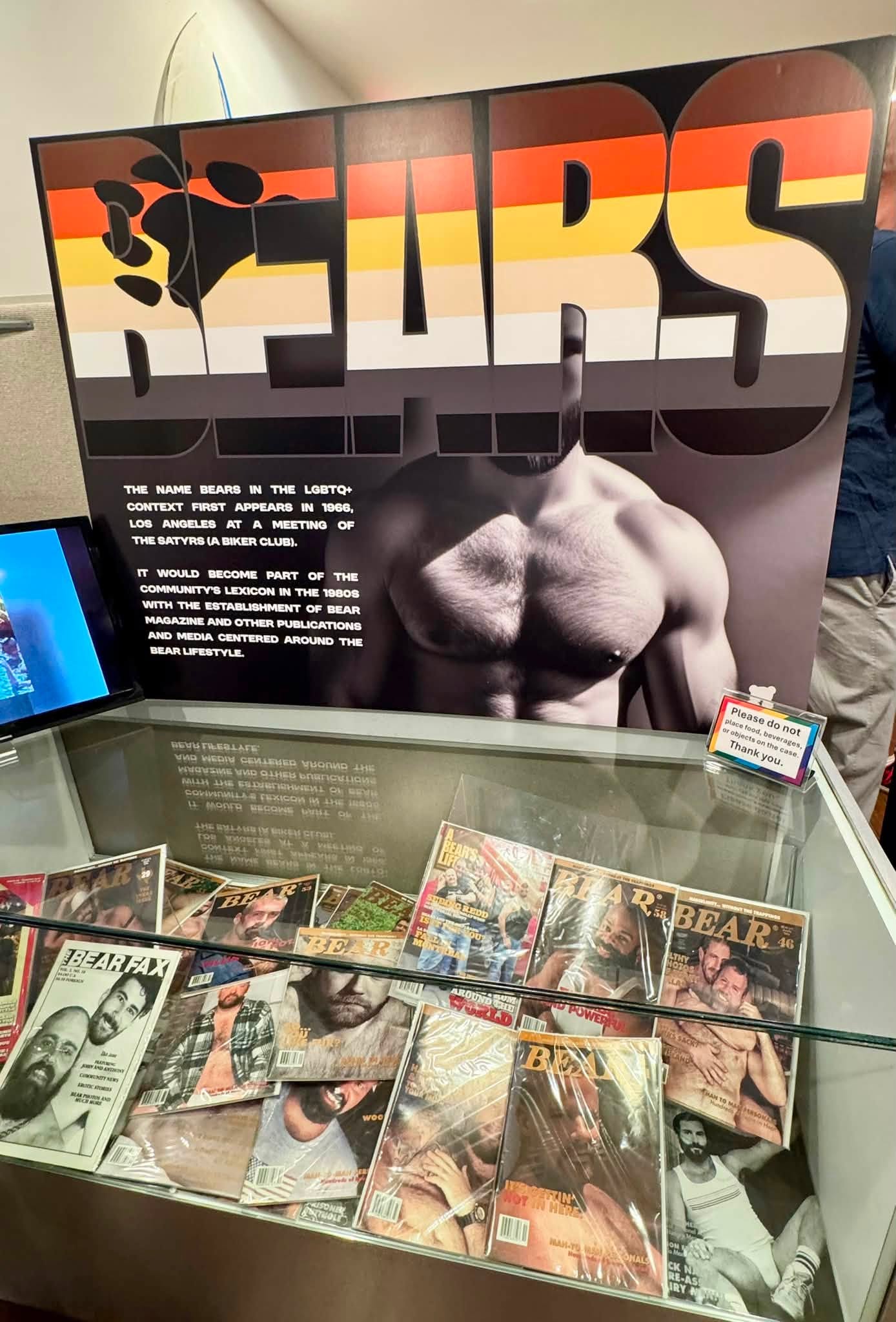 Large sign reading BEARS with rainbow-orange gradient, overlaid on a muscular black-and-white torso image in a display window of vintage bear magazines below.