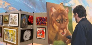 Artist in a dark shirt painting a large lion portrait on an easel, as colorful artwork fills a display wall behind him.