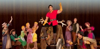 Theater scene: a male lead in a red tunic leaps on stage in skates while a colorful ensemble of dancers applauds around him.