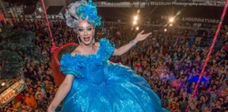Drag queen in a voluminous bright blue gown with matching headpiece entertaining a packed crowd from a float above the audience.