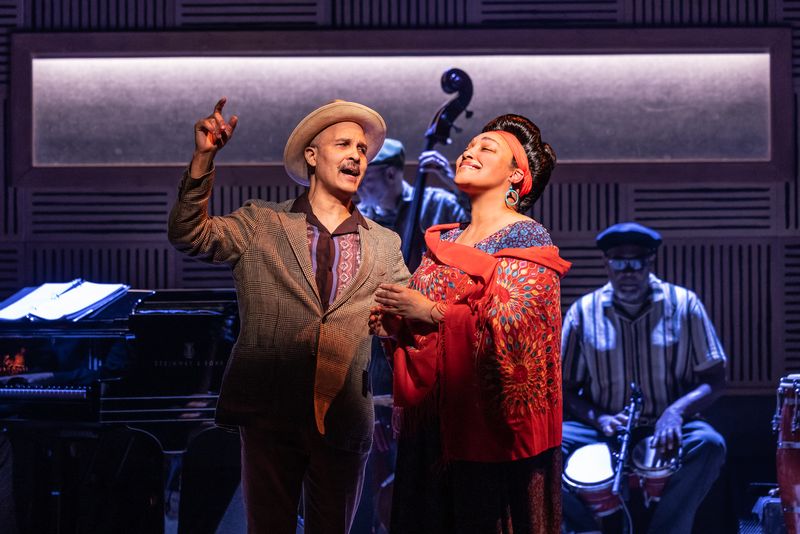 Two performers sing together on a retro jazz stage, a man in a hat and a woman in a red shawl, with a live band behind them.