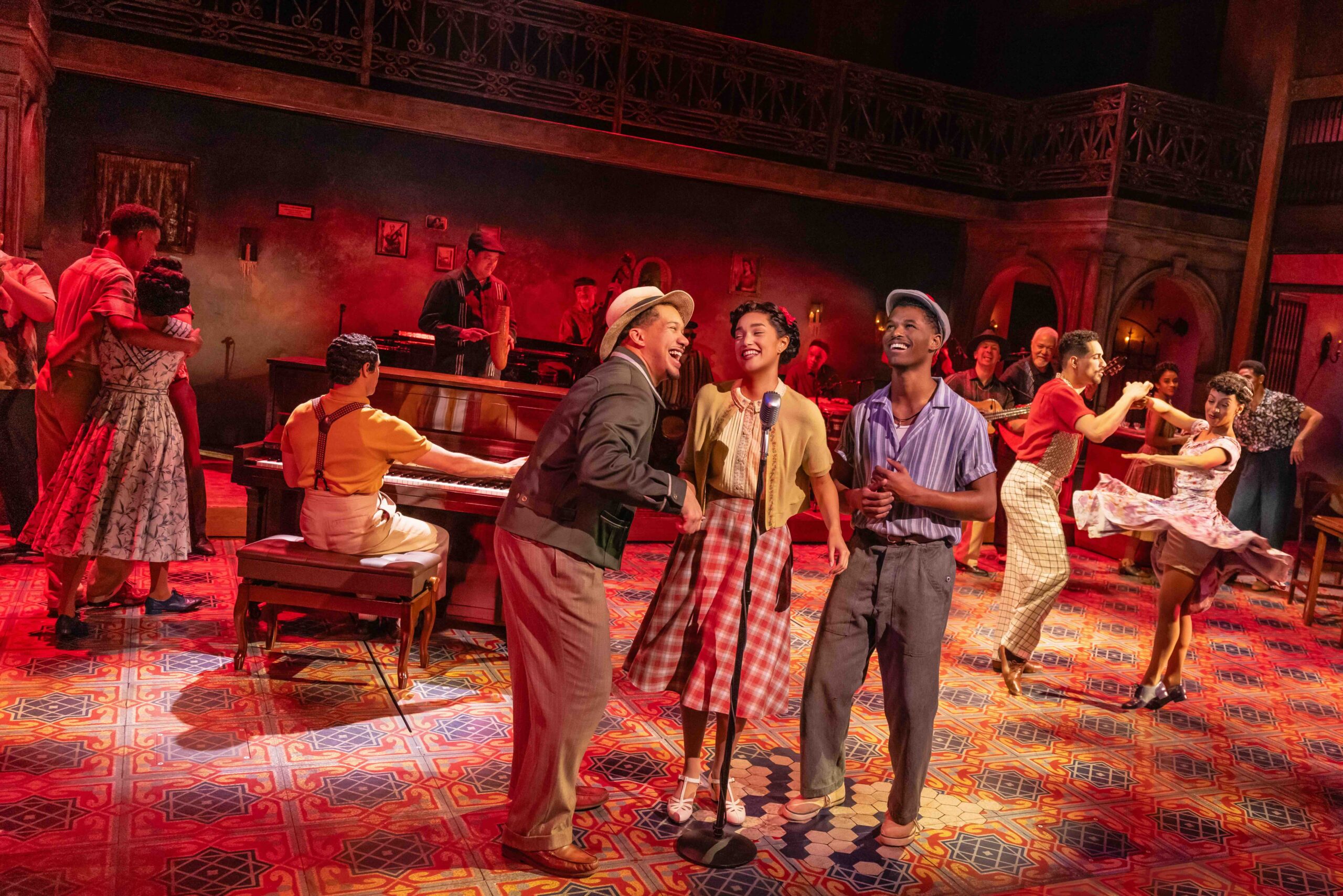 Dancers and musicians perform on a vintage-style stage; a singer at a microphone smiles center stage with others dancing and playing instruments in the background.