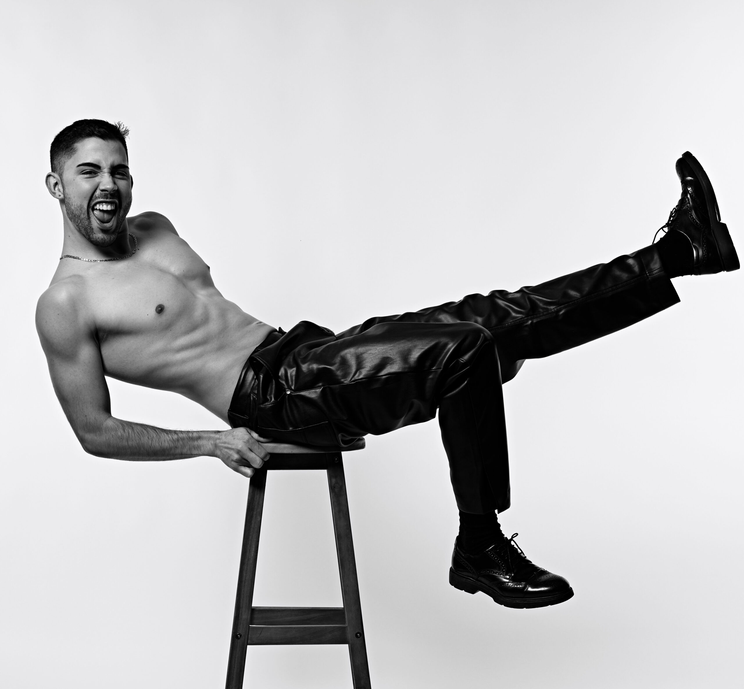 Black-and-white photo of a shirtless man on a ladder, kicking his leg forward in black leather pants and boots, energetic pose.