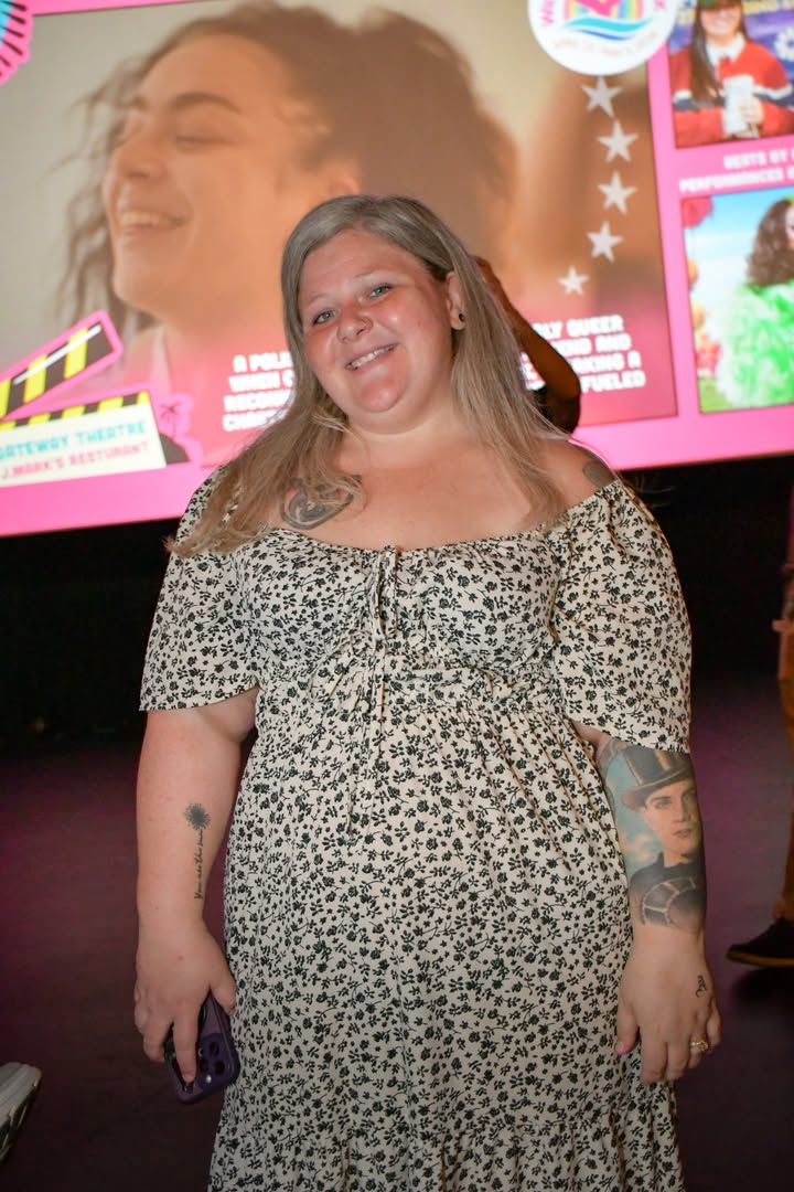 Outshine Film Festival’s Fall Edition – Fort Lauderdale and Miami from Oct 15-25 | Hotspots Magazine Hotspots Magazine Smiling woman with gray hair in a floral off-shoulder dress, holding a purple phone, standing in front of a bright presentation screen.