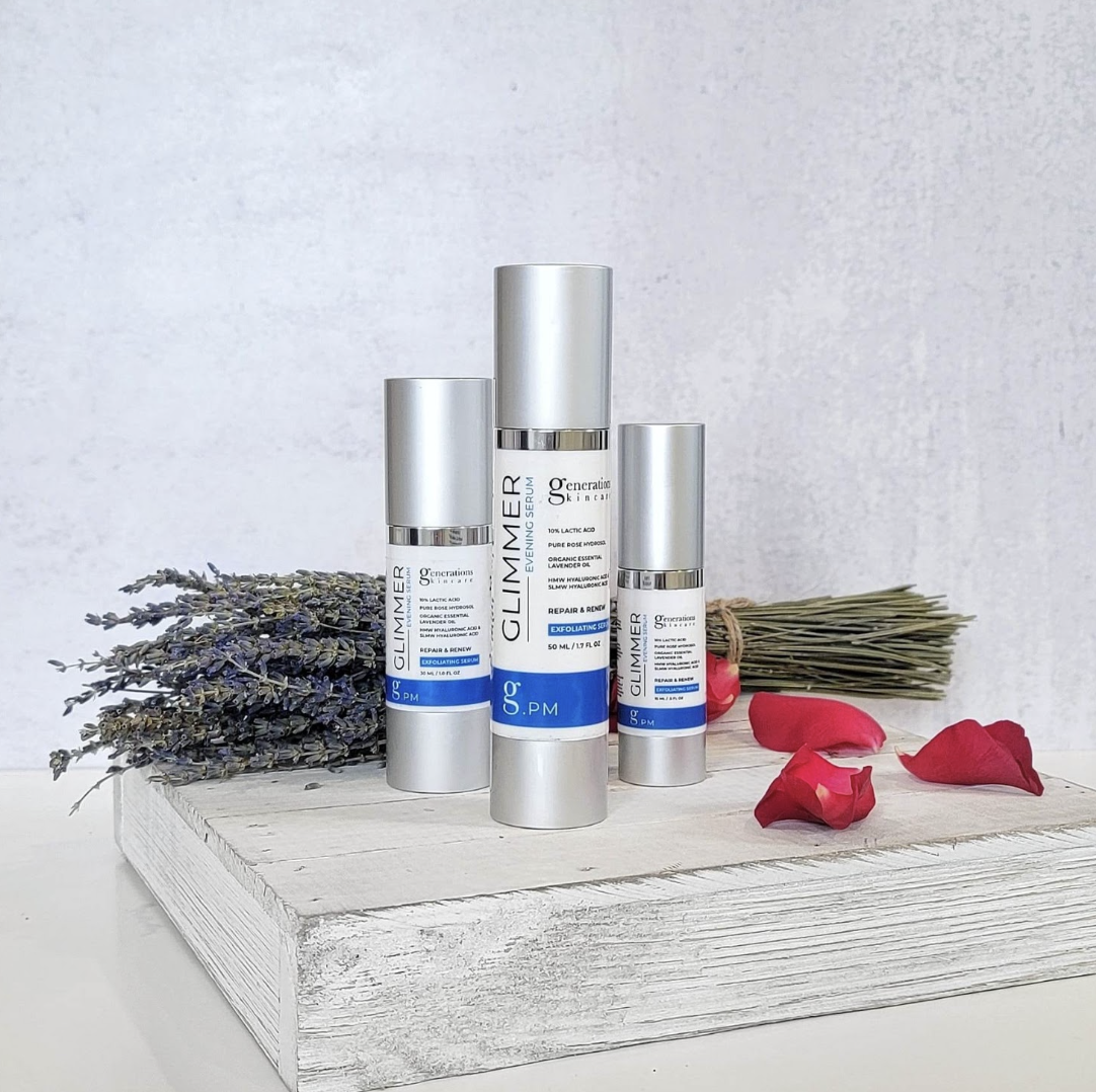 Three silver cosmetic bottles labeled Glimmer g.PM on a white wooden block with lavender bouquet and rose petals nearby.