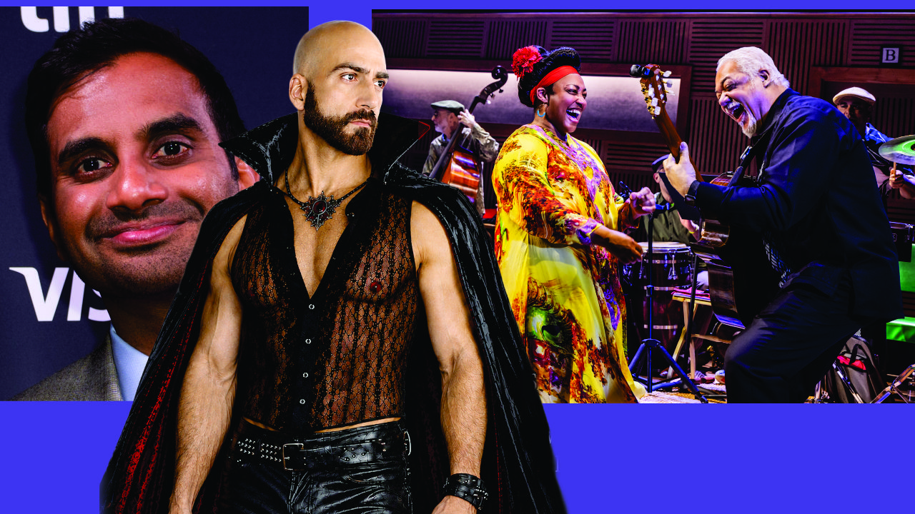 Person in a sheer black lace shirt and cape stands in the foreground as a lively band performs behind him on a colorful stage backdrop.